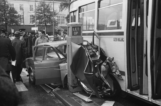 Schwarzes und weißes Bild eines Bus, der auf der Seite der Straße in ein Auto gekracht ist, mit Menschen und Bäumen im Hintergrund.