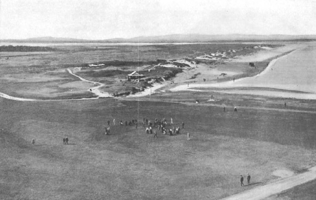 Schwarz-weiß-Foto von Golfern auf dem 18. Loch des Royal Birkham Golf Clubs, mit saftig grünem Rasen, verstreuten Häusern, sanften Hügeln und einem hellblauen Himmel im Hintergrund.