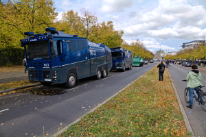 Eine Gruppe von Polizeifahrzeugen, die am Straßenrand geparkt sind, mit einer Person, die auf einem Fahrrad auf der rechten Seite fährt, viele Menschen, die Fahrräder halten, Gras und trockene Blätter auf dem Boden, Bäume, Gebäude und ein bewölkter Himmel im Hintergrund.