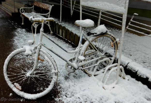 Ein Fahrrad auf einer schneebedeckten Straße mit einer nahen Bank, Geländern, einem Gebäude im Hintergrund und Text unten.