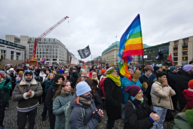 Eine große Gruppe von Menschen steht vor einem Gebäude und hält Fahnen und Schilder mit Text, darunter "Lgbtq+ Rechte Demonstration in Berlin", einige tragen Mützen und Taschen, mit Gebäuden, einem Kran und einem bewölkten Himmel im Hintergrund.