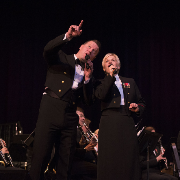 Ein Mann und eine Frau in Fracks, die Mikrofone halten, auf einer Bühne mit einer Sitzreihe von Musikern dahinter in einer dunklen Umgebung.