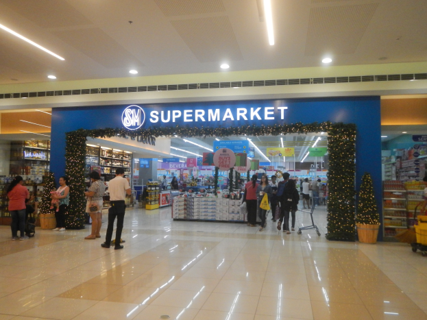 Eine Gruppe von Menschen, die vor einem Supermarkt stehen, einige halten Einkaufstaschen, mit einem Einkaufswagen, Topfpflanze, Regalen mit Gegenständen, Schildern und Deckenbeleuchtung im Hintergrund.