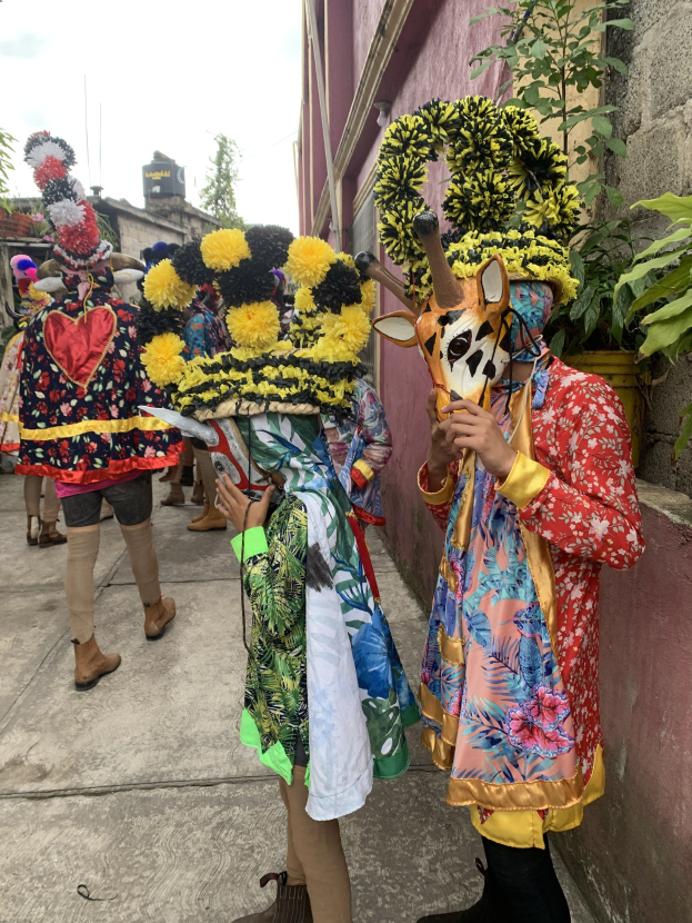 Eine Gruppe von Menschen in bunten Kostümen mit verschiedenen Gegenständen, die vor einem Gebäude mit Bäumen und einem klaren blauen Himmel stehen, wahrscheinlich an einem Faschingsumzug teilnehmend.