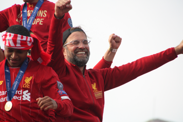 Liverpool-Trainer Jurgen Klopp feiert mit seinen Spielern den Sieg in der UEFA Champions League, während zwei medaillentragende Personen in der Nähe stehen und der Himmel im Hintergrund zu sehen ist.