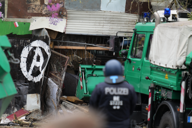 Ein Polizeibeamter steht vor einem mit Graffiti beschmierten Gebäude, umgeben von Fahrzeugen, Bäumen und anderen Gegenständen, mit einem menschlichen Gesicht am unteren Rand des Bildes.
