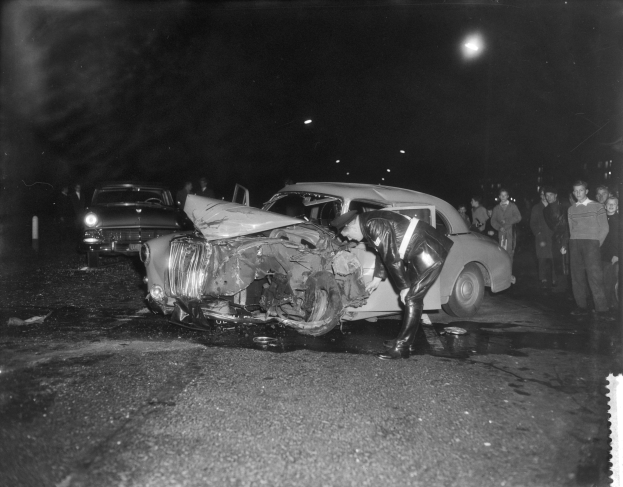 Schwarzes und weißes Foto eines auf der Straße gestrandeten Autos mit mehreren Menschen in der Nähe und beleuchteter Hintergrundbeleuchtung.