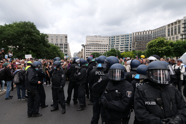 Eine große Gruppe von Polizisten steht vor einer Menge auf einer Straße, die von Bäumen und Gebäuden gesäumt ist, unter einem bewölkten Himmel, wobei einige Demonstranten Kameras halten.