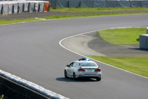 Ein Polizeiauto fährt auf einer Rennstrecke mit Gras auf beiden Seiten, mit Menschen, Zelten und anderen Gegenständen im Hintergrund.