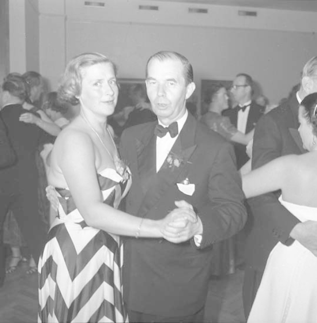 Ein Mann in einem schwarzen Blazer, weißem Hemd und schwarzen Hosen tanzt mit einer Frau in einem unbekannten Outfit bei einem formellen Ereignis, indem sie sich an den Händen halten und zur Musik schaukeln, vor einer Wand als Hintergrund.