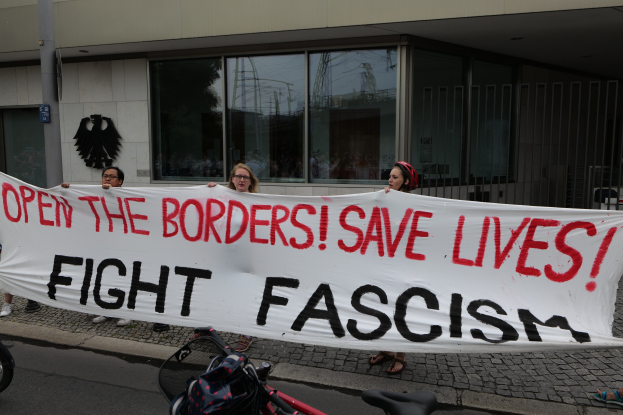 Gruppe von Menschen mit einem Transparent "Öffnet die Grenzen, rettet Leben, kämpft gegen Faschismus" vor einem Gebäude mit Glasfenstern und einem Pfahl, mit einem Fahrrad und einer Tasche auf der Straße.