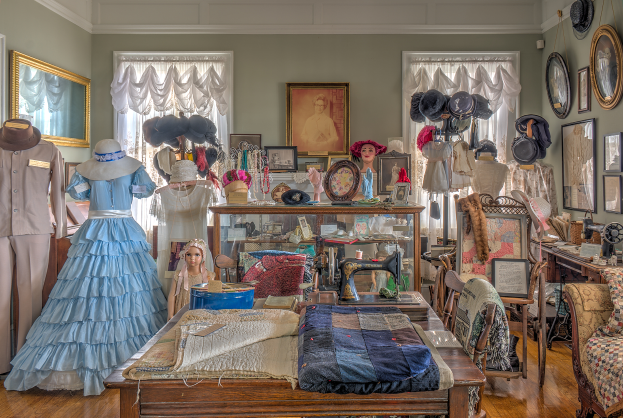 Ein Raum im Museum of Fine Arts in St. Louis, Missouri, gef├╝llt mit Puppen in verschiedenen Outfits, einem Tisch mit Klamotten und einer N├Ąhmaschine, St├╝hlen mit Kissen, einem Schrank mit Gegenst├Ąnden, Fotorahmen an den W├Ąnden, Fenstern mit Vorh├Ąngen und einer Decke.