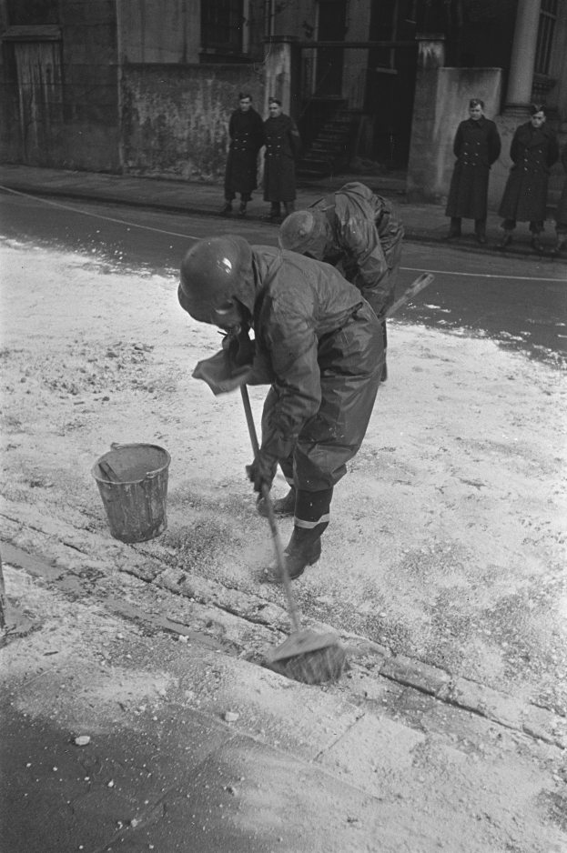 Ein Mann in einem Helm und Handschuhen fegt Schnee vom Gehweg mit einem Besen, während eine Gruppe von Menschen auf dem Bürgersteig in der Nähe steht, mit einem Gebäude mit Fenstern und einer Tür im Hintergrund.