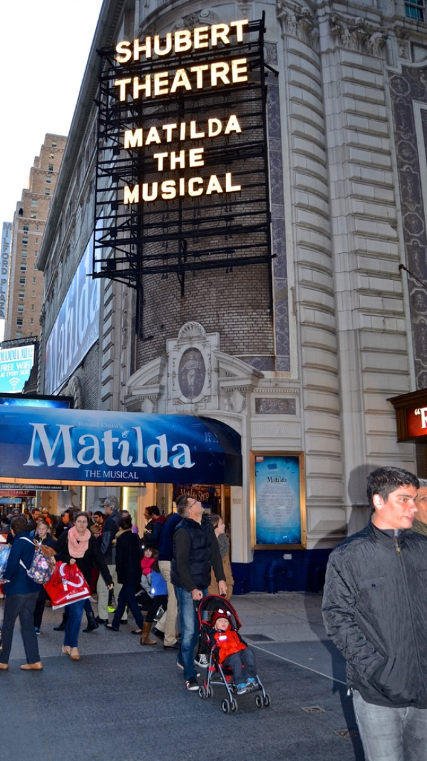 Eine Gruppe von Menschen steht in der Nähe von Gebäuden und Werbetafeln, mit einem Baby im Kinderwagen.
