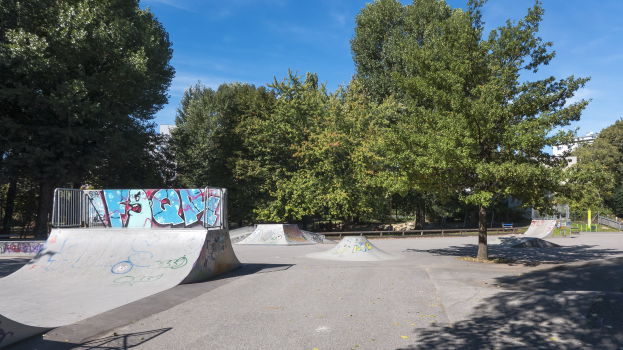 Ein Park mit einer zentralen Skaterrampe, umgeben von Bänken, Geländern, Gras, Pflanzen, Bäumen und Gebäuden im Hintergrund.