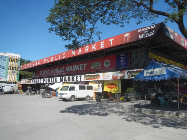 Öffentlicher Markt mit einem weißen Van vorne geparkt, umgeben von Gebäuden, Geschäften, Schildern, Fahrzeugen auf der Straße, Menschen unter einem Zelt sitzend, Geländern, Bäumen und einem klaren blauen Himmel.
