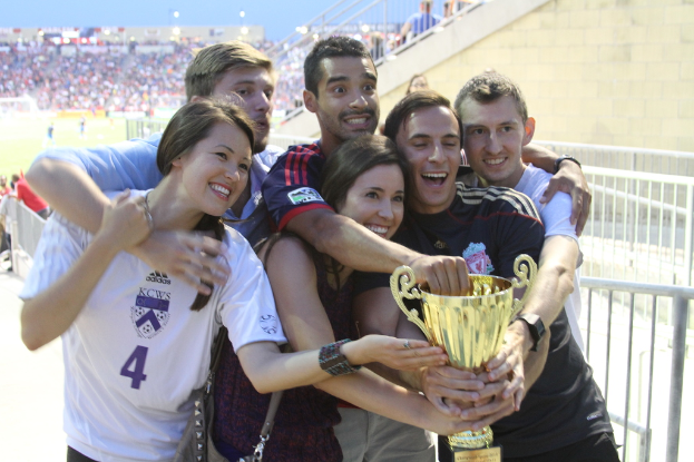 Sechs Personen, zwei Frauen und vier Männer, halten einen Pokal und lächeln, posieren für ein Foto vor einem Eisenzaun, mit einer Wand und einem Zaun oben rechts, Menschen und Lichter im Hintergrund, möglicherweise in einem Stadion.