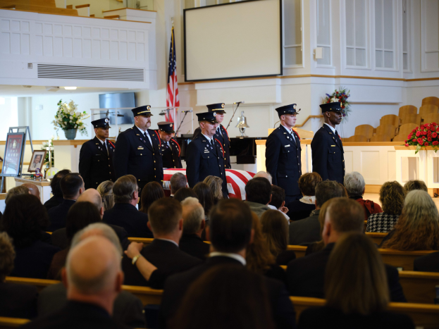 Eine Gruppe uniformierter Personen mit Mützen vor sitzenden Trauernden bei einer Beerdigungszeremonie, mit Blumensträußen, einer Flagge, einem Bildschirm und einer Wand im Hintergrund.