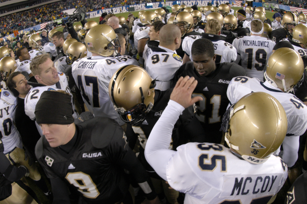 Eine Gruppe von Fussballspielern in Helmen versammelt sich auf einem Rasenfeld, mit einer Menschenmenge auf den Tribünen und Transparenten mit Text im Hintergrund.