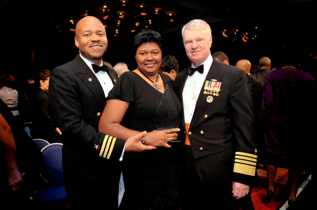 Zwei Männer und eine Frau auf einem roten Teppich; die Frau hält eine Trophäe, alle drei sind formell gekleidet, mit anderen Menschen und beleuchteter Decke im Hintergrund.