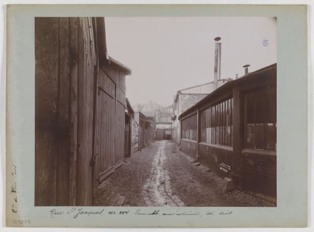 Ein altes Schwarz-Weiß-Foto einer Schotterstraße mit Gebäuden auf beiden Seiten und Text am unteren Bildrand.