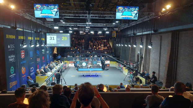 Eine große Menschenmenge sitzt vor einer Bühne mit einem Tischtennis-Tisch in der Mitte, Stühlen auf der linken Seite, einer Wand auf der rechten Seite und Deckenlampen und Fernseher im Hintergrund bei der 2019 Jumbo-Weltmeisterschaft.