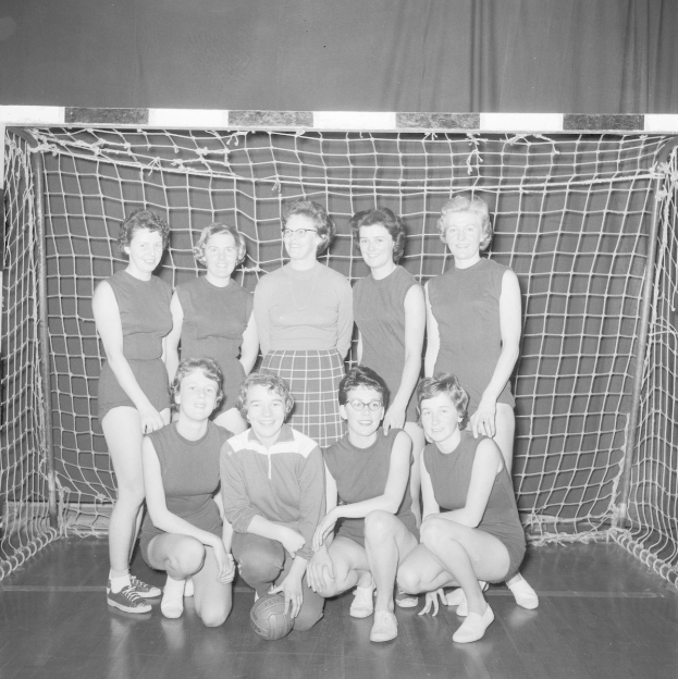 Schwarzes und weißes Foto einer Gruppe von Frauen, die vor einem Fußballtor posieren, mit einem Ball auf dem Boden und einem Vorhang als Hintergrund.