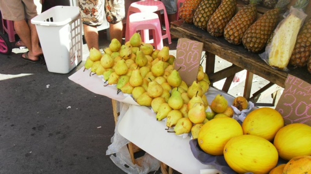 Eine Marktszene mit verschiedenen Fruächten auf einem Gestell, ein paar Höcker, einige Menschen und zwei Preisplakate.