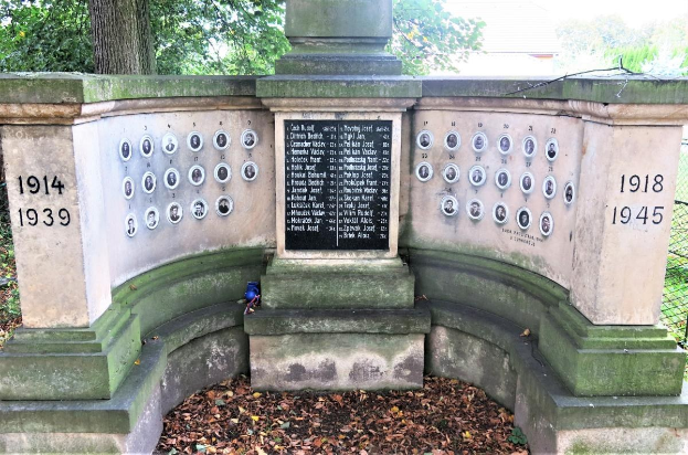 Ein Holocaust-Denkmal-Monument in einem jüdischen Friedhof in Berlin, das eine Wand mit Text und Zahlen zeigt, umgeben von Bäumen und einem Zaun mit verstreuten trockenen Blättern.