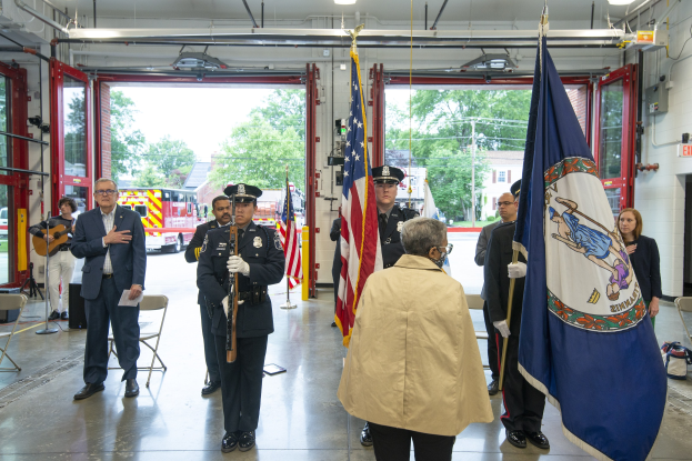 Eine Gruppe von Mitgliedern der New York City Fire Department steht vor einem Feuerwehrauto, einige halten Fahnen, mit verstreuten Stühlen, Taschen und anderen Gegenständen drumherum, unter Deckenlampen, mit Türen, Fahrzeugen, Bäumen, Gebäuden, Masten, Drähten und einem klaren blauen Himmel im Hintergrund.