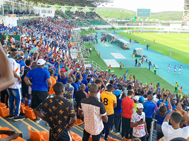 Eine große Menschenmenge versammelt sich vor einem eingezäunten Fußballfeld, einige sitzen und andere stehen, mit Blick auf Hügel unter einem klaren blauen Himmel während der African Cup of Nations 2019.