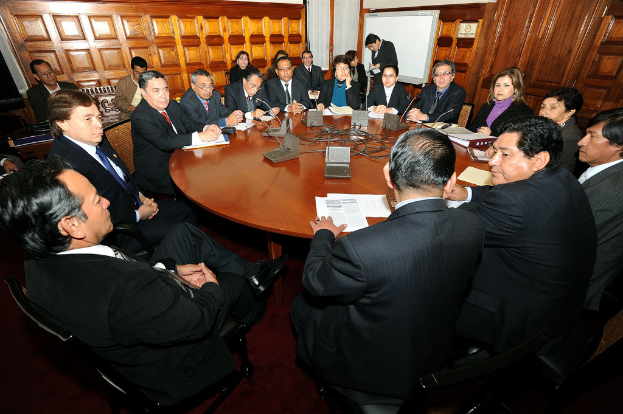 Rundtischgespräch mit Delegierten, die über Mikrofone, Papiere, Kabel und Dateien auf dem Tisch diskutieren, mit einer hölzernen Wand im Hintergrund.