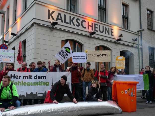 Eine Gruppe von Menschen mit Protestschildern und Plakaten vor einem Gebäude, mit zwei Personen im Vordergrund auf einem Gegenstand sitzend und einem Müllcontainer auf der rechten Seite, vor einem Hintergrund von Gebäuden mit Fenstern und Schildern in Deutschland.