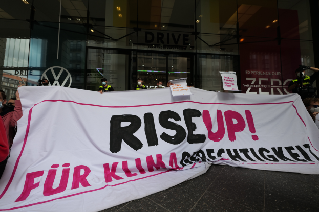 Eine Gruppe maskierter Personen mit einem "Steh Auf"-Schild und Schildern vor einem hell erleuchteten Gebäude mit Glaswänden und -türen.