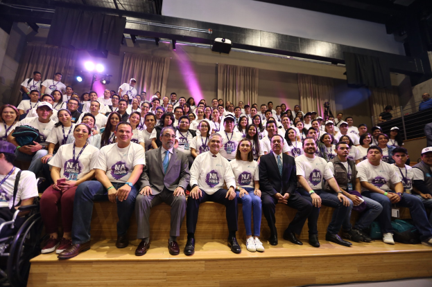 Eine Gruppe von Menschen sitzt auf einer Bühne, mit einer Person im Rollstuhl links vor Vorhängen, Scheinwerfern und einer Wand auf einer NAWBC-Konferenz.