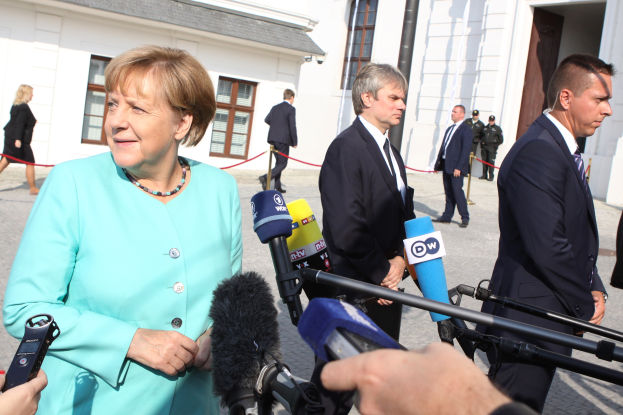 Bundeskanzlerin Angela Merkel spricht vor der Reichstag in Berlin mit Reportern, umgeben von einer Gruppe von Menschen, einige halten Mikrofone, mit Gebäuden im Hintergrund.