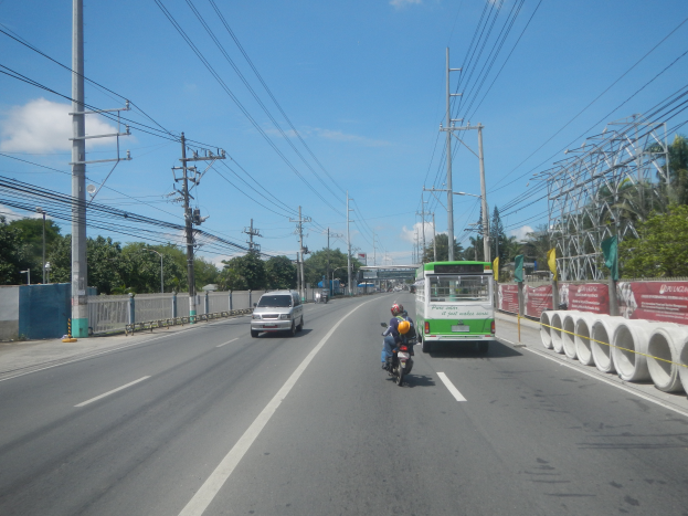 Eine Person fährt mit einem Motorrad auf einer Straße mit Strommasten und Drähten, neben einem Bus mit weißen Rohren, einer roten Fahne, einer Begrenzungsmauer, Bäumen und Laternen, unter einem klaren blauen Himmel.