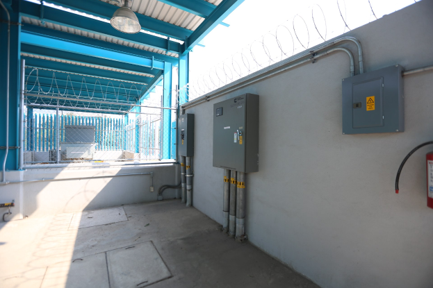 Ein Raum mit einem Feuerlöscher auf der rechten Seite, elektrischen Schächten und Rohren an der Wand, einer Deckenleuchte und einem Zaun im Hintergrund, der an eine elektrische Unterstation erinnert.