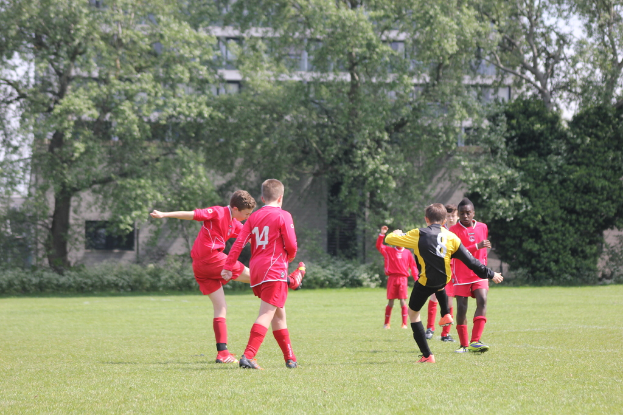 Fünf Jungs in roten T-Shirts und Shorts spielen Fußball auf einem grünen Feld mit Bäumen und Gebäuden im Hintergrund.