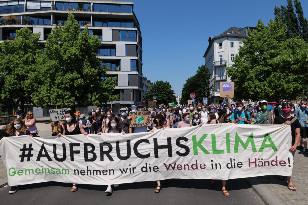 Maskentragende Menschen halten ein Banner mit der Aufschrift "Aufbruchsklima" vor einem Gebäude mit Bäumen und einem klaren Himmel.