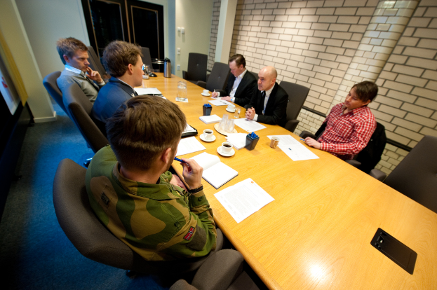 Eine Gruppe von Menschen sitzt um einen Tisch mit Tassen, Büchern, Papieren, einer Flasche, einem Krug und Stiften.