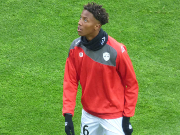 Ein junger Mann in roter Jacke und weißen Shorts steht auf einem Fußballfeld und schaut rechts mit entschlossener Miene, trägt Baseballhandschuhe.