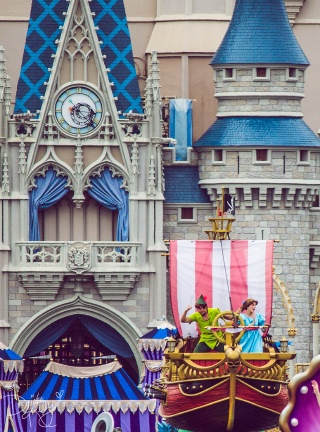 Eine bootähnliche Struktur auf der rechten Seite mit tanzenden Menschen und einer Flagge, mit einem Gebäude mit Uhr und Vorhängen im Hintergrund.