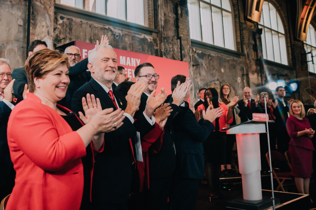 Eine Gruppe von Menschen, wahrscheinlich Liberale, steht vor einem Publikum, klatscht feierlich, mit einem Podium, Mikrofon und Texttafel rechts, Stühlen, einer Fahne, einer Wand mit Fenstern und Lichtern im Hintergrund.