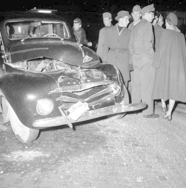 Schwarz-weißes Bild eines schwer beschädigten Autos am Straßenrand mit eingedellter Motorhaube und zersplitterten Fenstern, umgeben von einer Gruppe von Menschen, die es besorgt untersuchen.