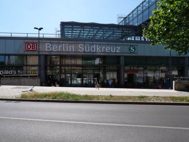 Großes, gläsernes Gebäude des Berliner Südkreuz-Bahnhofs mit sichtbarem Text, umgeben von Straßeninfrastruktur, Fahrzeugen, Fußgängern, Fahrrädern, Bäumen und einem klaren Himmel.