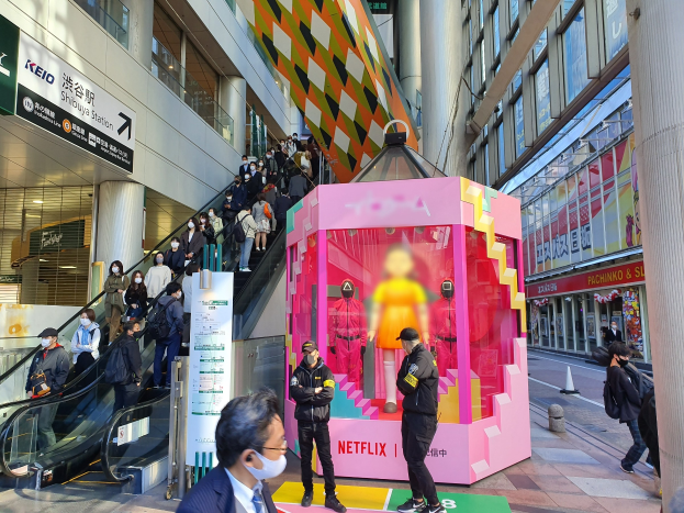 Eine Gruppe von Menschen steht vor einem Gebäude mit Glaswänden und Texttafeln, einige tragen Masken, während eine Rolltreppe mit Fahrgästen zu sehen ist; ein pinker Kasten in der Mitte zeigt Schaufensterpuppen und trägt die Aufschrift "Netflix's new Netflix booth in Tokyo."