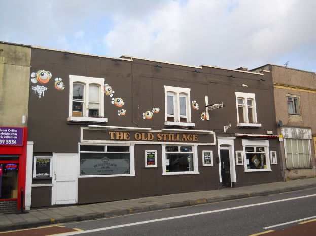 Eine Straßenszene außerhalb der Stadt mit einer Straße, einem Gehweg und Gebäuden mit sichtbaren Türen und Fenstern, darunter eines mit der Aufschrift "The old stillage"; eine rote Wand und Glastür links, ein violettes Schild mit Text und ein klarer Himmel darüber.