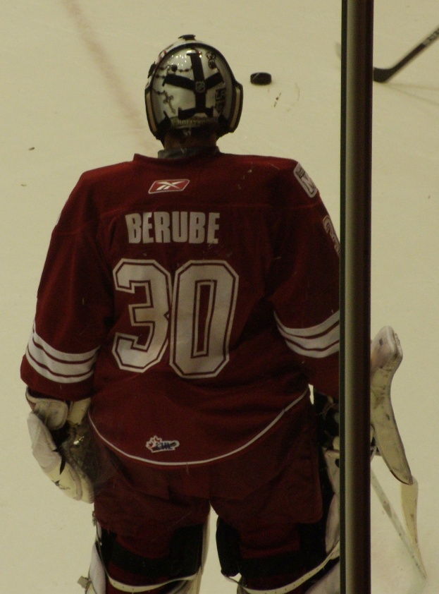Ein Hockey-Spieler in Helm, Handschuhen und einer nummerierten Jersey steht auf dem Eis vor einem Tor mit einem Pfosten rechts daneben.