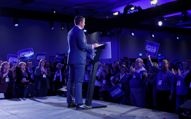 Ein Mann steht an einem Rednerpult mit einem Mikrofon und spricht zu einer Menge, während einige Leute klatschen und Schilder halten, unter Deckenleuchten auf einer Wahlveranstaltung.